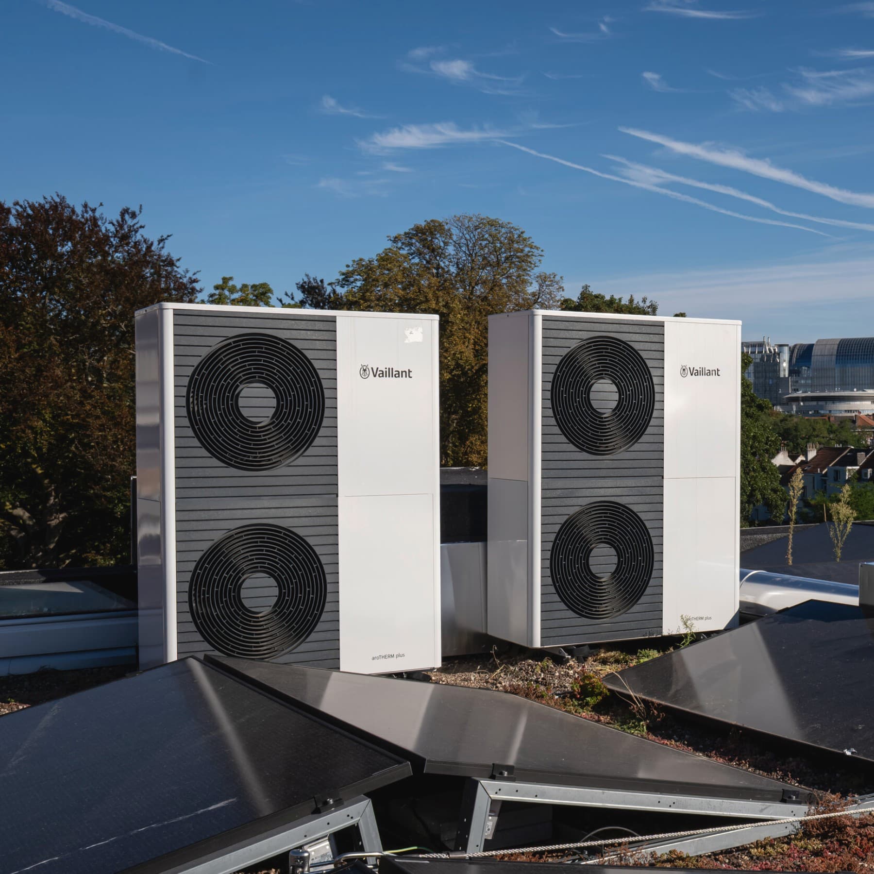 Twee dubbele warmtepompen op een dak, voor de buitenunits liggen zonnepanelen en op de achtergrond zie je bomen