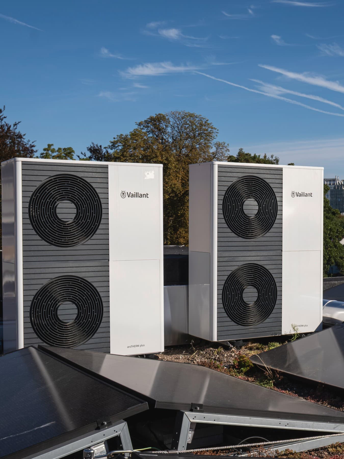 Twee dubbele warmtepompen op een dak, voor de buitenunits liggen zonnepanelen en op de achtergrond zie je bomen