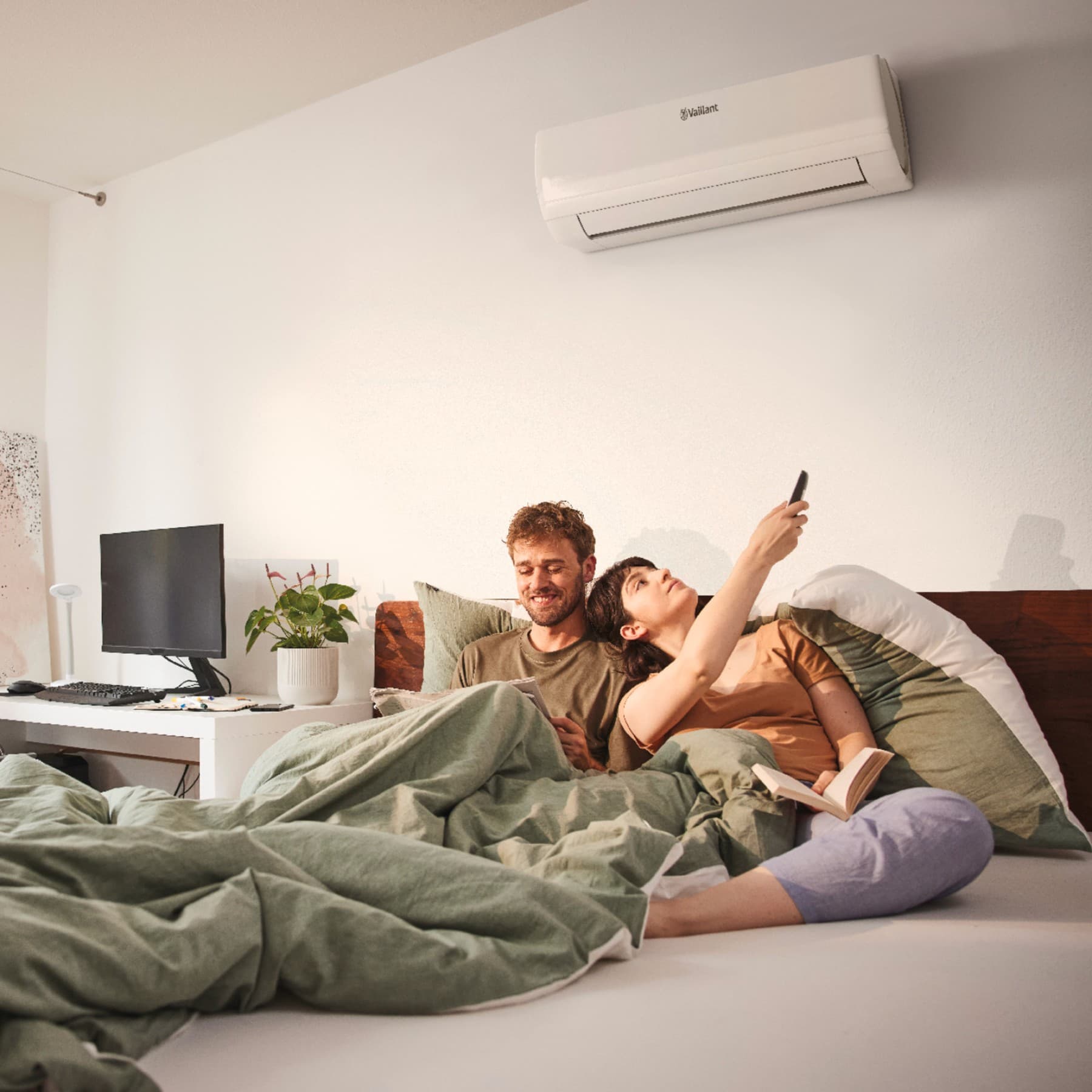 Koppel ligt nog in bed onder groene dekens, de vrouw zet met een afstandsbediening de airco aan. Op de achtergrond staat een bureau met een plant en scherm met toetsenbord