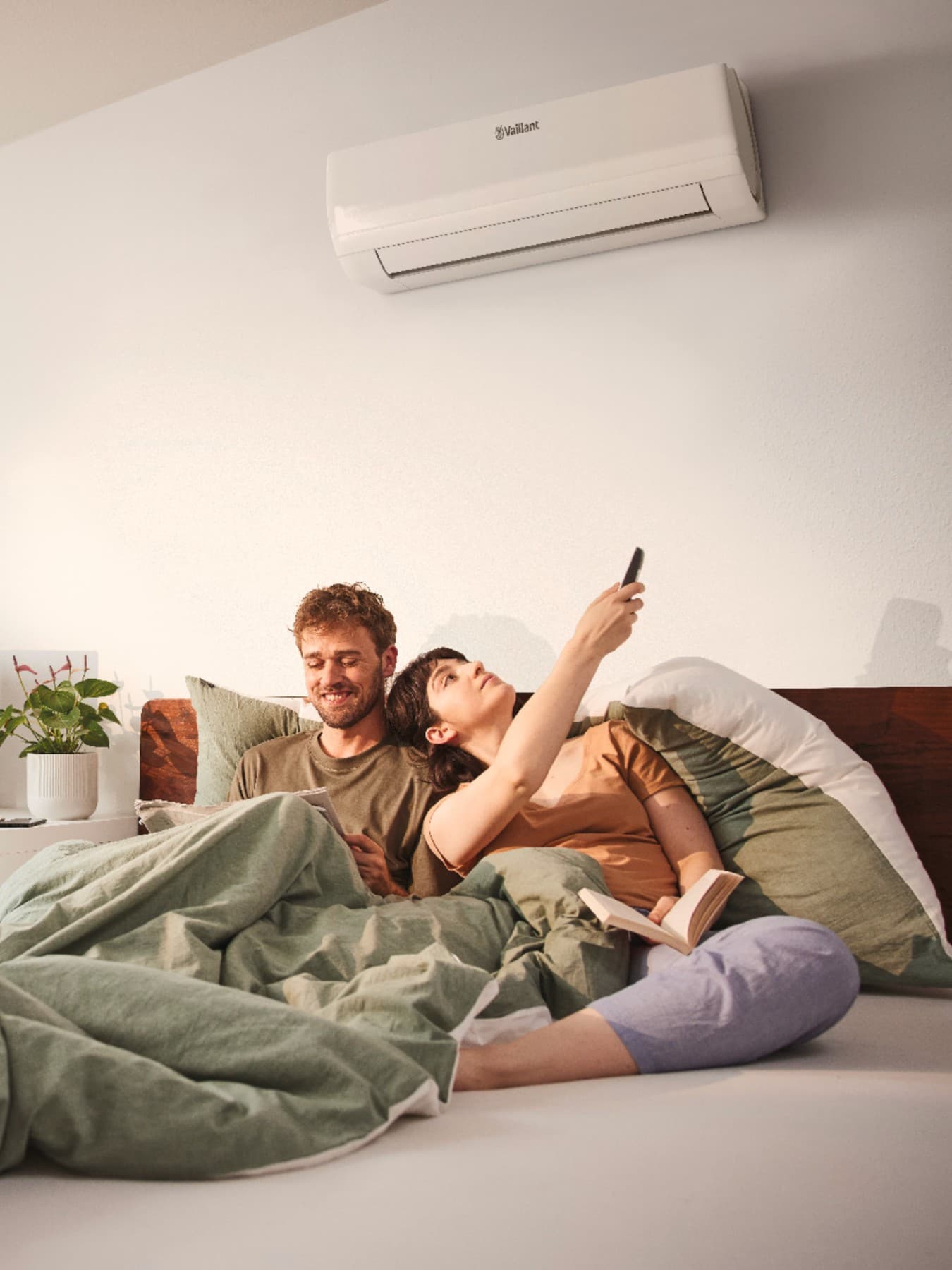 Koppel ligt nog in bed onder groene dekens, de vrouw zet met een afstandsbediening de airco aan. Op de achtergrond staat een bureau met een plant en scherm met toetsenbord