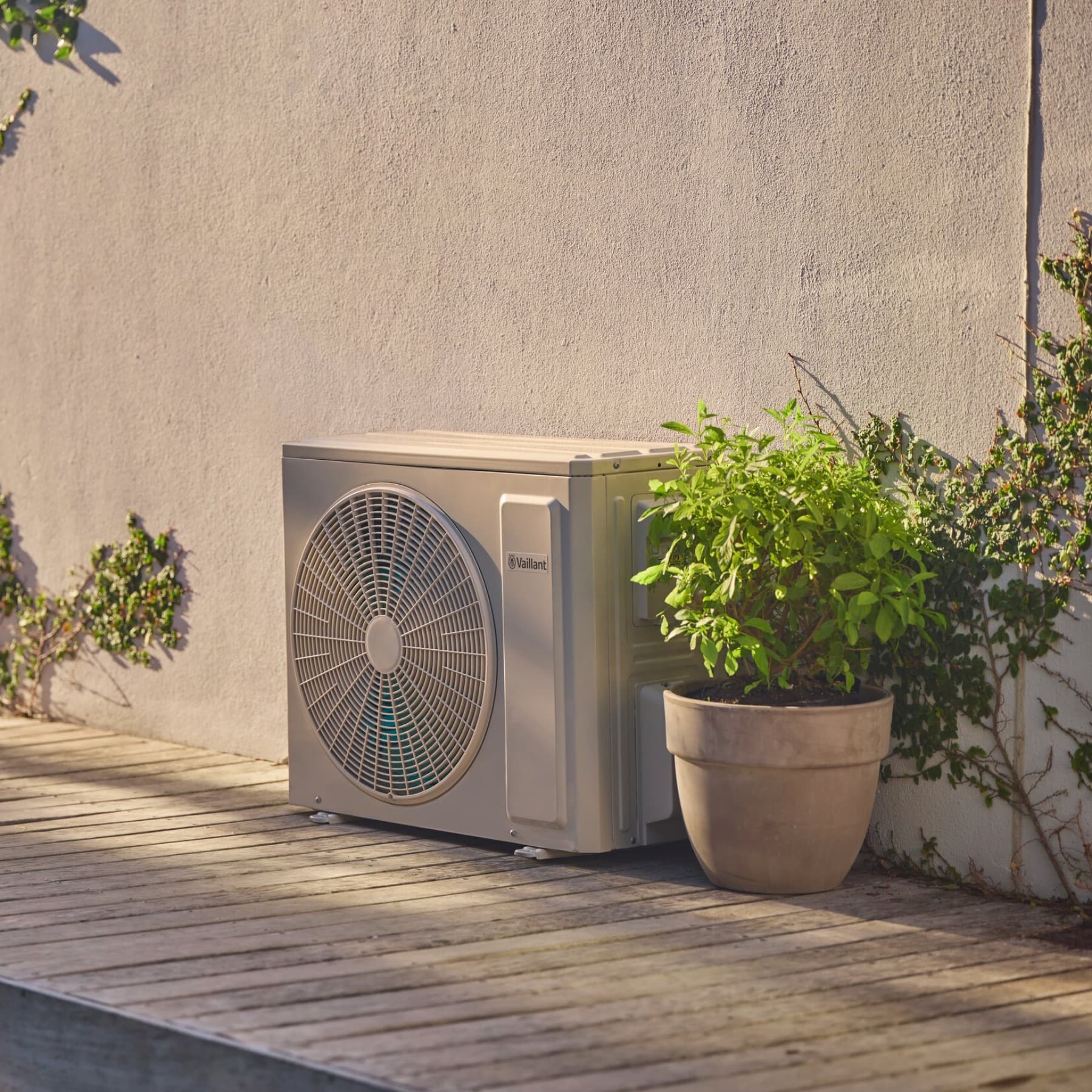 Buitenunit van een airco naast een grijze gevel omringd door planten