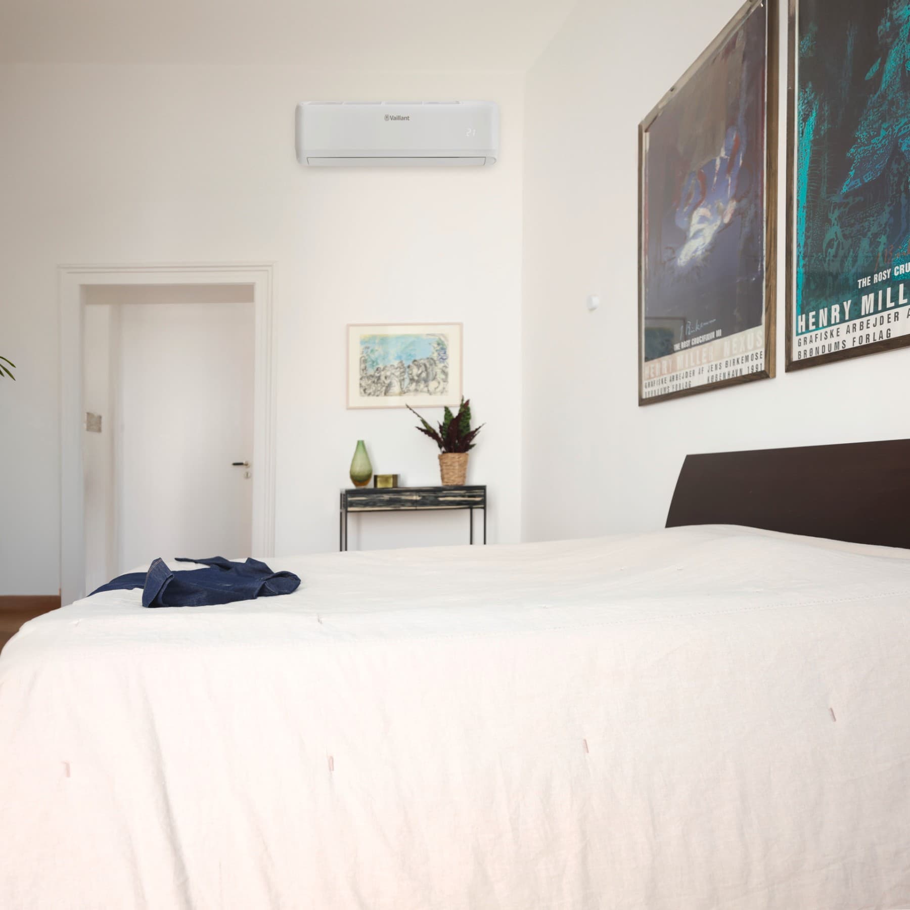 Kamer waar een blauw shirt op een bed ligt, er hangen kaders aan de muur en er staat een tafeltje met een plant en vaas op
