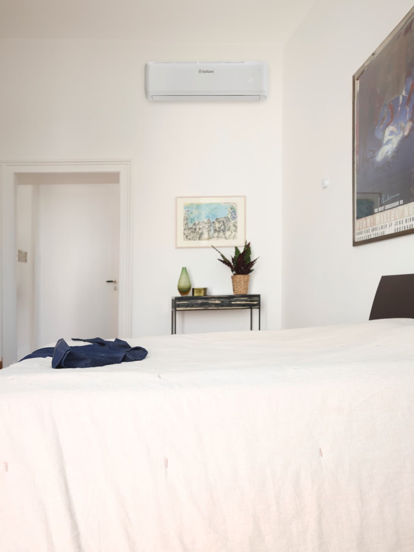 Kamer waar een blauw shirt op een bed ligt, er hangen kaders aan de muur en er staat een tafeltje met een plant en vaas op