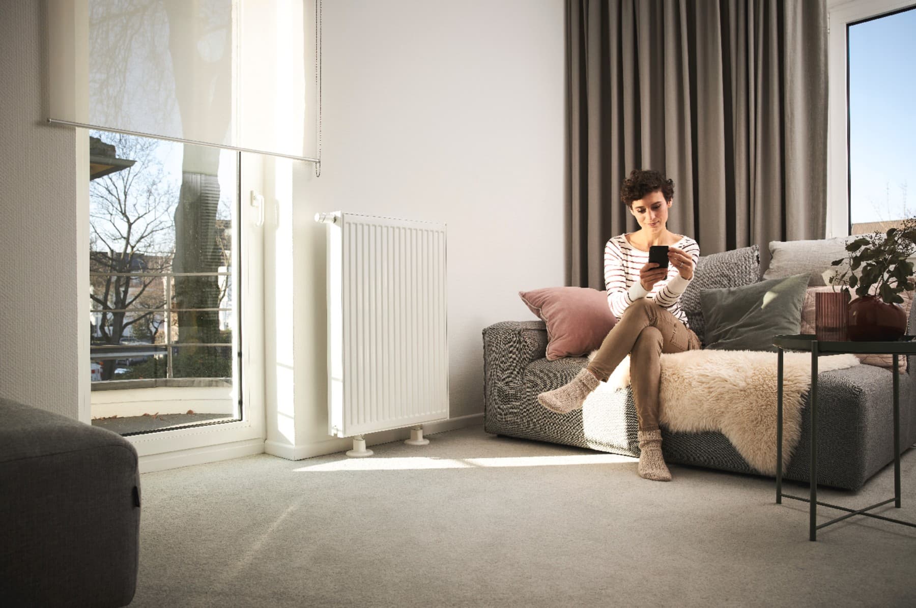Vrouw zit op bank naast radiator en kijkt naar mobiele telefoon
