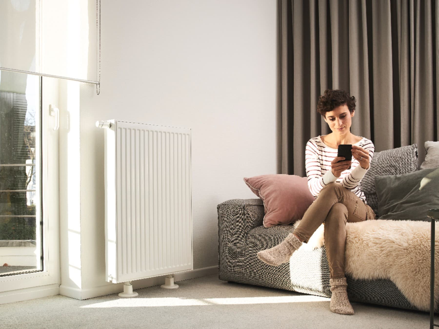 Vrouw zit op de bank en gebruikt een smartphone. De bank staat voor een raam en naast een radiator.