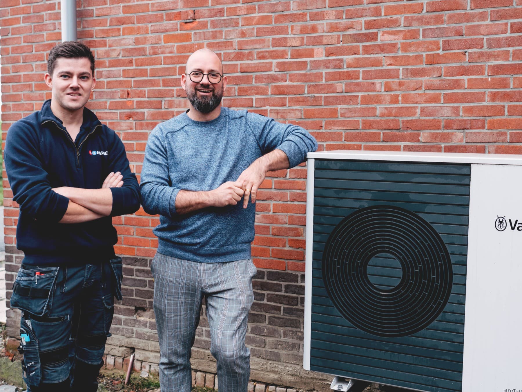 Installateur en klant naast buitenunit van een Vaillant warmtepomp met rode bakstenen muur op de achtergrond