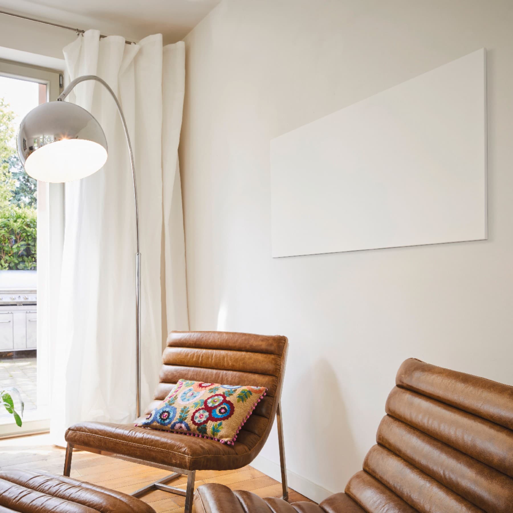 Bruinlederen stoelen met felgekleurd kussen naast een zilverkleurige ronde lamp aan een raam met witte gordijnen en een wandpaneel aan de witte muur