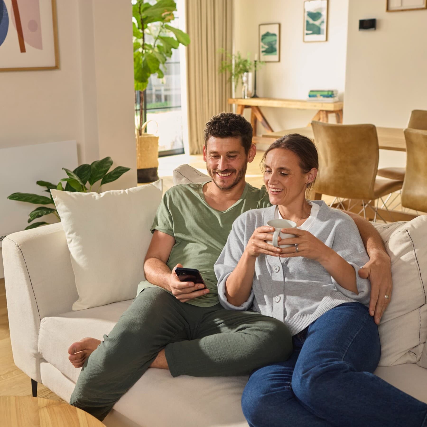 Een koppel zit op de bank en kijkt naar een smartphone. Op de achtergrond is de Vaillant aroVAIR plus ventilatorconvectorconsole te zien.