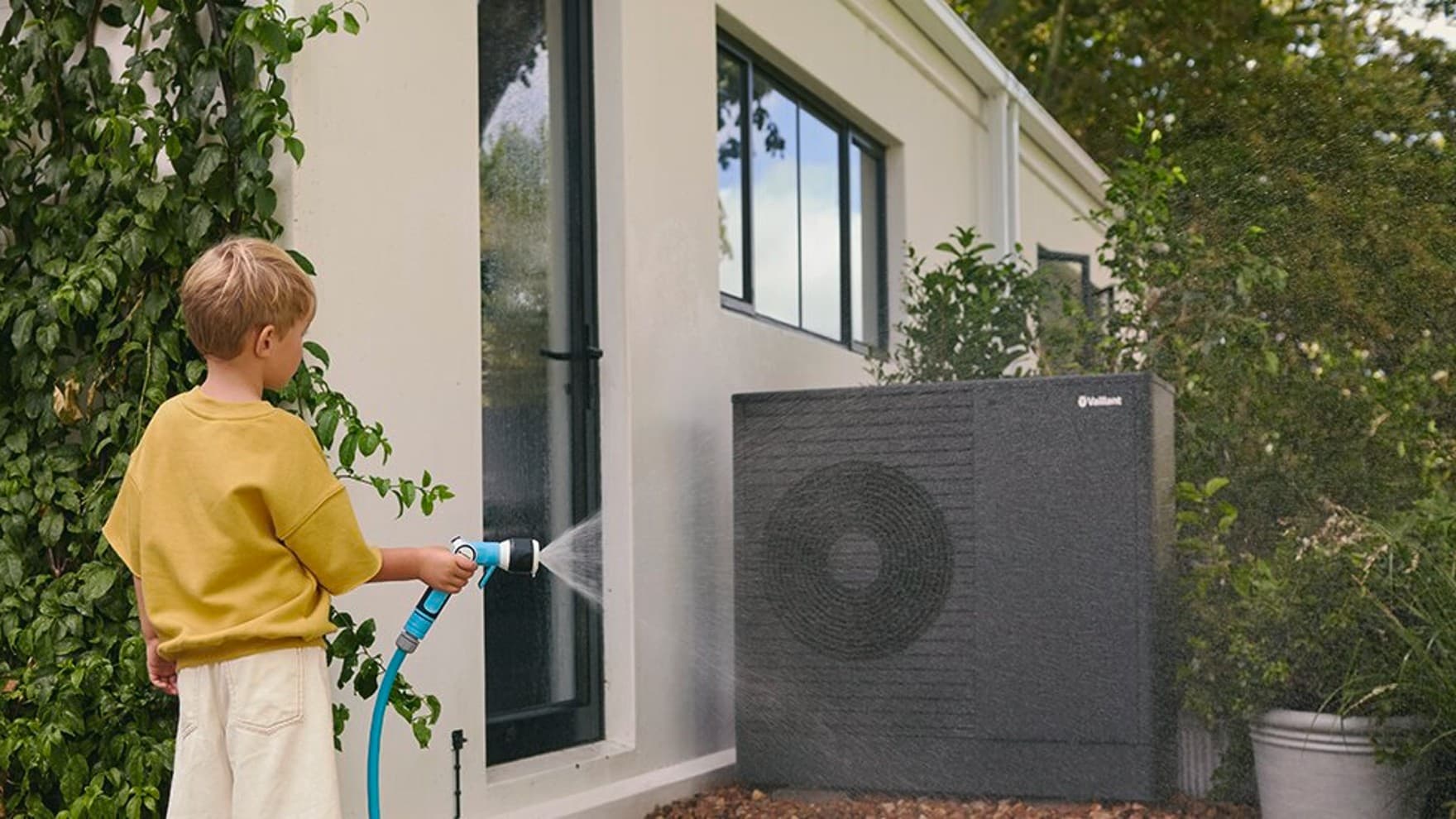Een jongen sproeit water op de warmtepomp met de tuinslang.