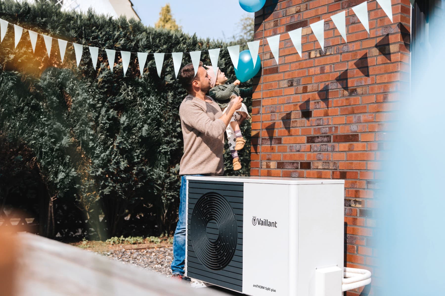 Man staat naast en gevel en houdt een kind in de lucht. Daarvoor staat een warmtepomp en er hangen vlaggen aan de muur