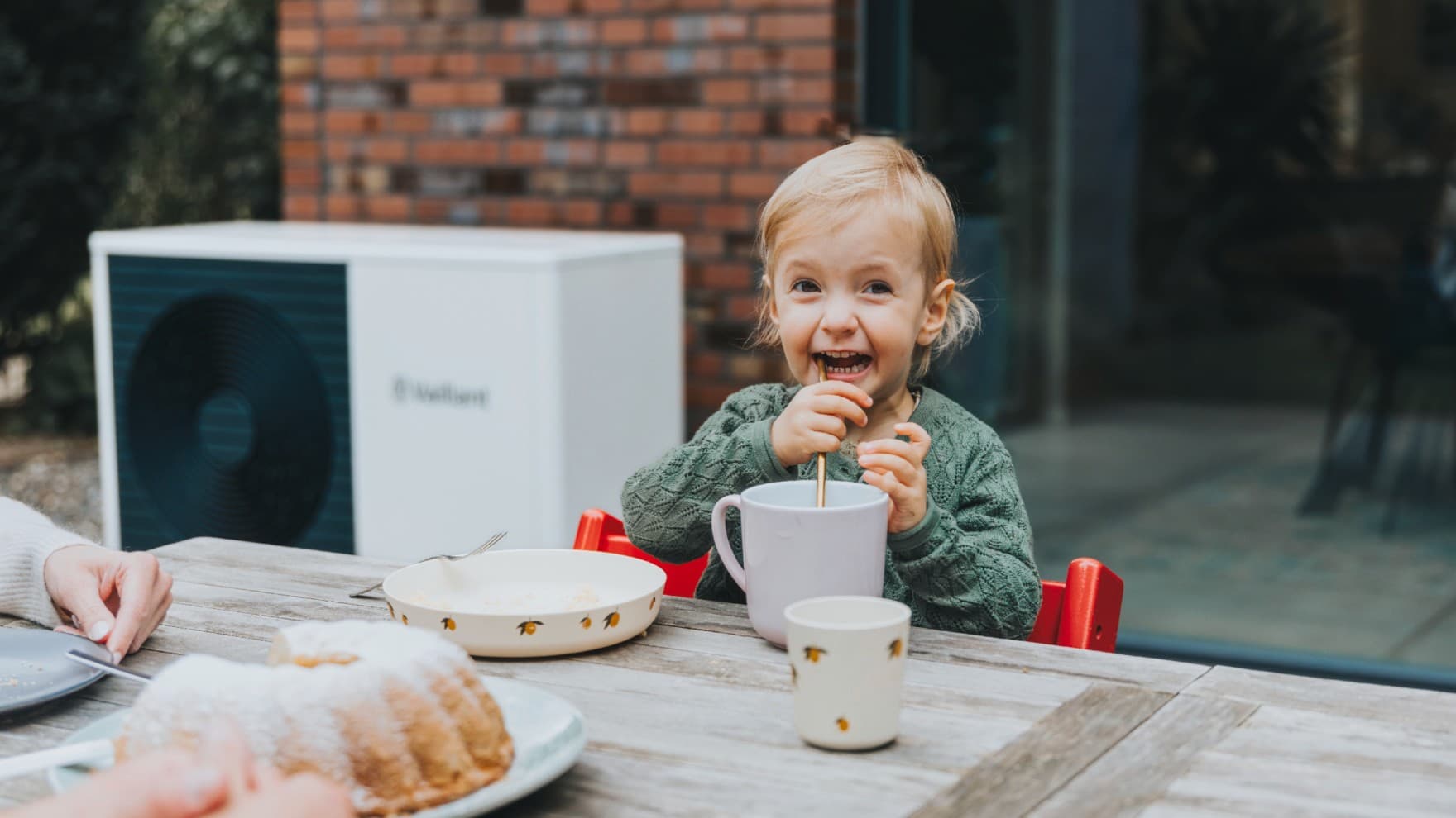 Kind drinkt met een rietje uit een tas. Op de achtergrond zie je een witte warmtepomp.