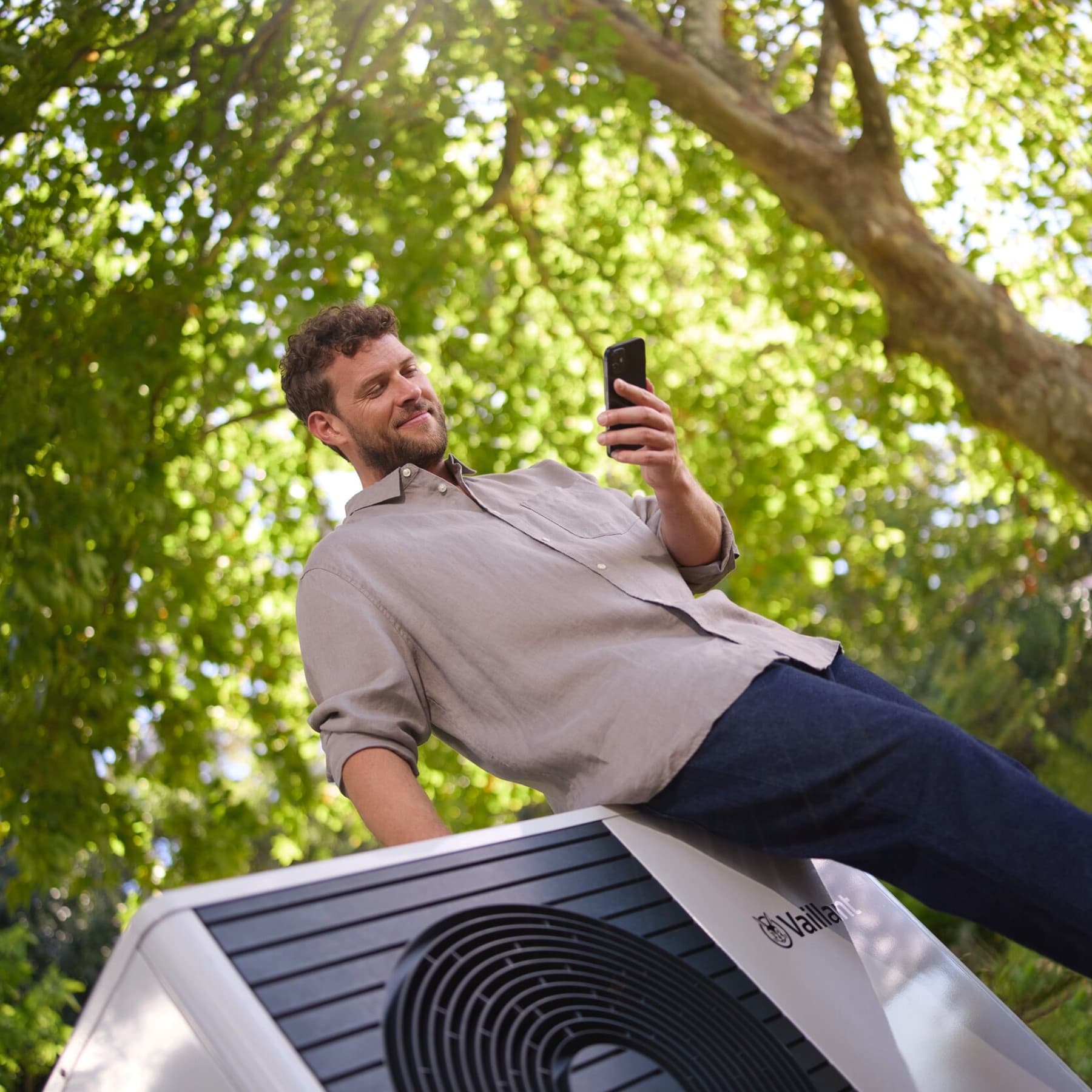 Man kijkt op zijn smartphone terwijl hij tegen een witte warmtepomp leunt. Op de achtergrond zie je de bladeren van een boom