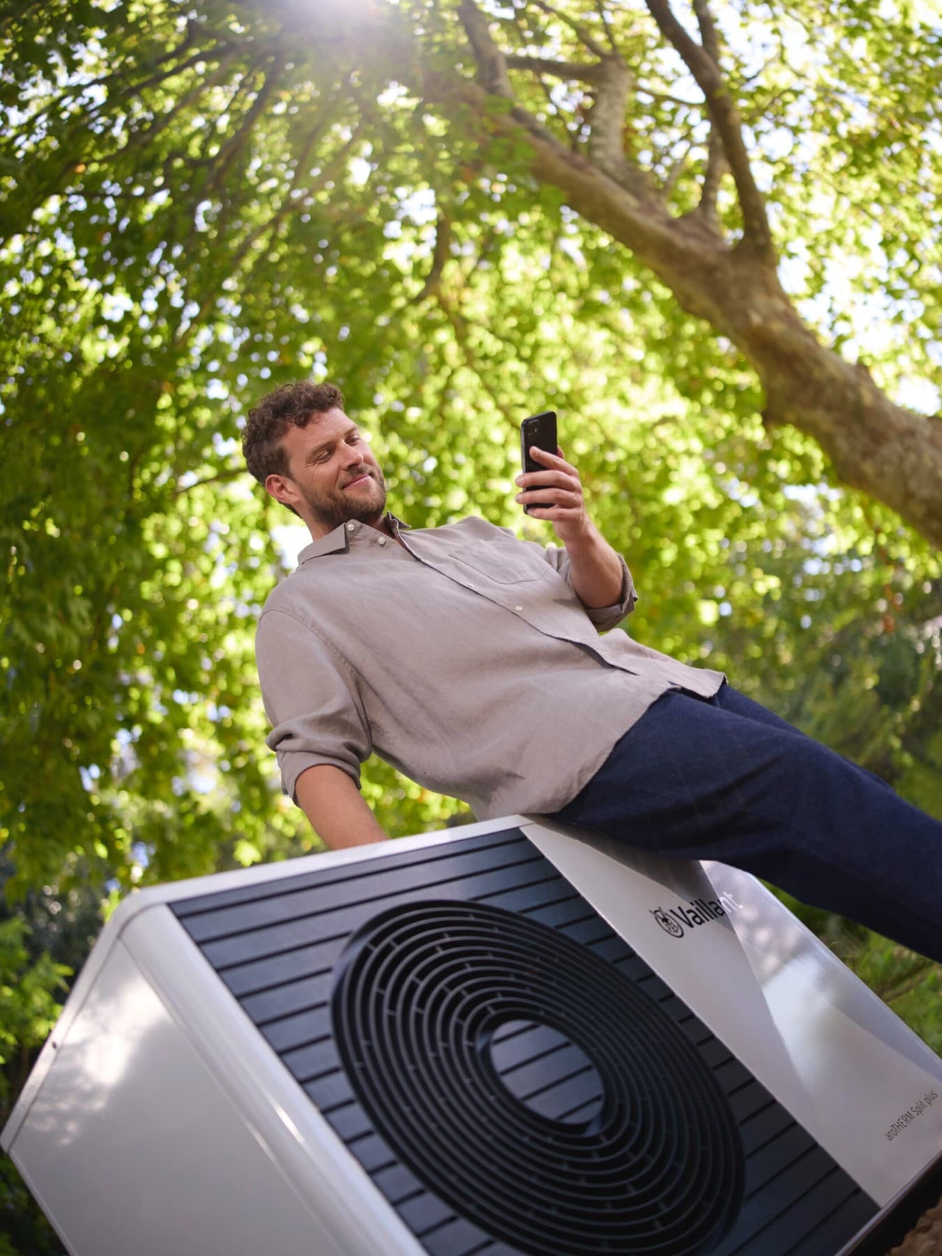 Man kijkt op zijn smartphone terwijl hij tegen een witte warmtepomp leunt. Op de achtergrond zie je de bladeren van een boom