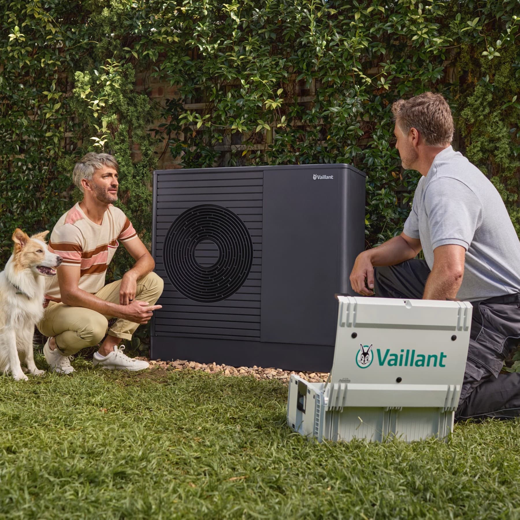 Installateur en klant bij een Vaillant‑warmtepomp in de tuin, met een hond.