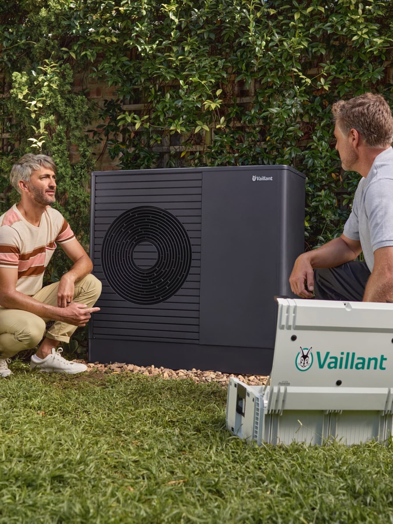 Installateur en klant bij een Vaillant‑warmtepomp in de tuin, met een hond.