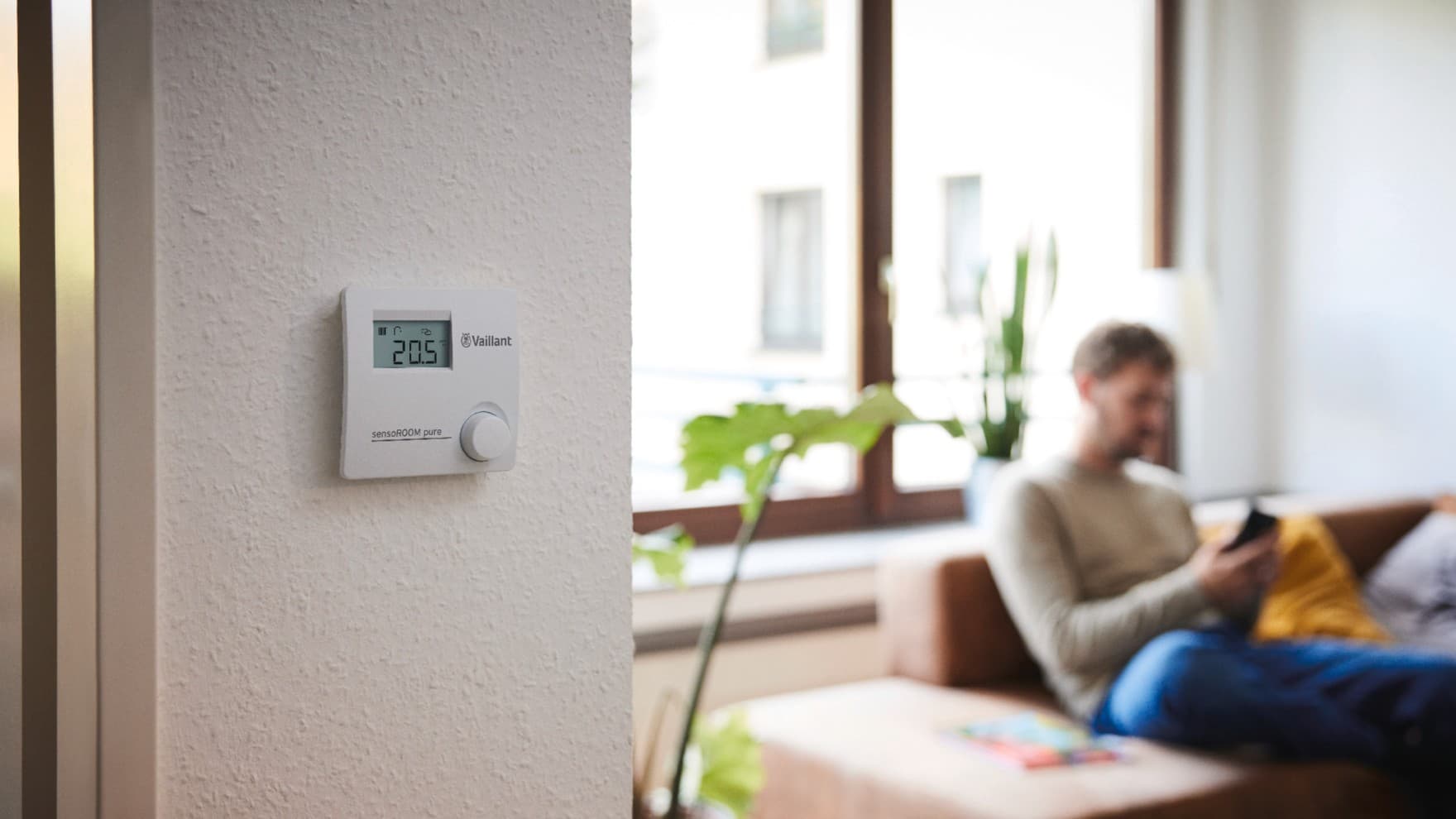 sensoROOM pure modulerende kamerthermostaat aan een muur, op de achtergrond zit een man op de zetel op zijn telefoon