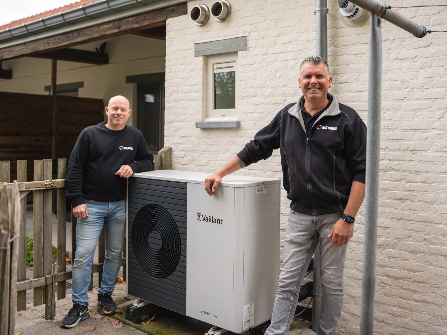 Twee mannen staan naast een witte warmtepomp in een tuin met op de achtergrond de gevel van een witte woning