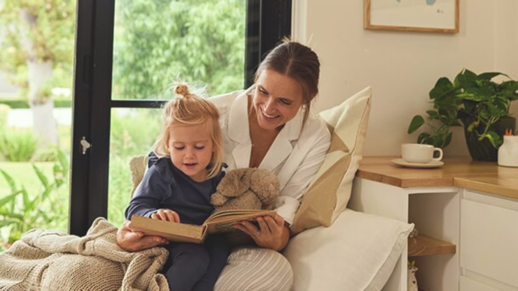 Moeder zit op een zetel en leest een boek voor aan een jong meisje. Ze houdt ook een beige pluchen konijn vast. Op de achtergrond staan een tas en een plant