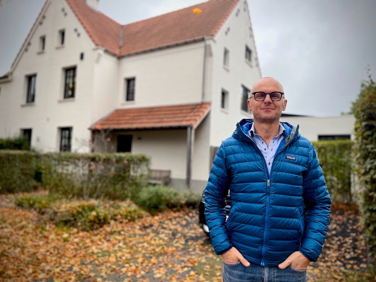 Man in blauwe jas staat voor een grote witte woning omringd door bomen